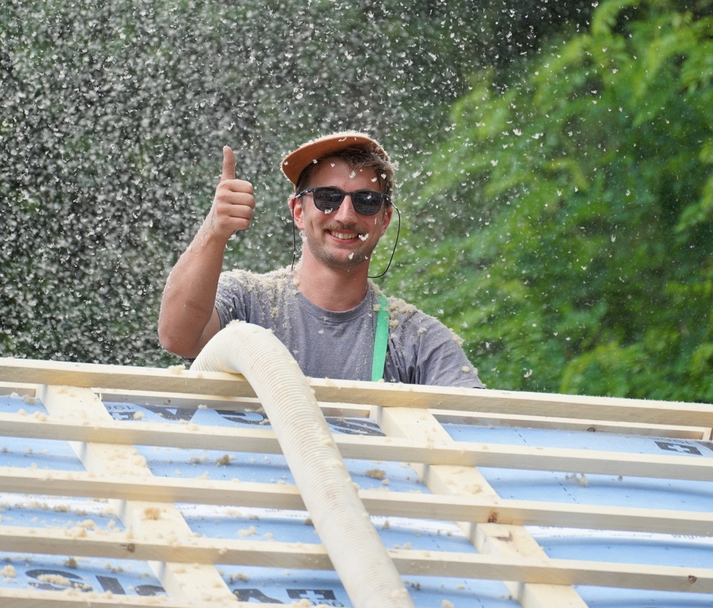 A smiling worker giving a thumbs up while installing insulation, with trees in the background